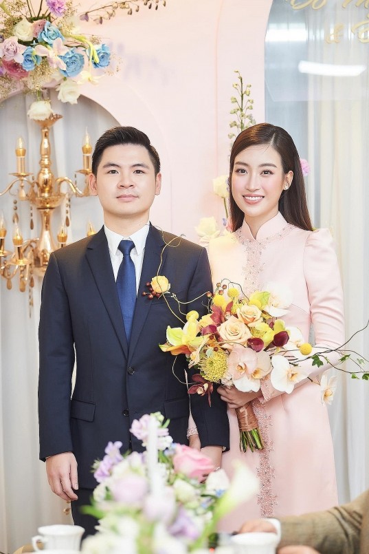 Đỗ Mỹ Linh diện áo dài hồng đính cườm dọc thân áo với điểm nhấn là phần cầu vai phồng sóng đôi bên ông xã. Đôi uyên ương nắm tay nhau rạng rỡ chào quan khách.